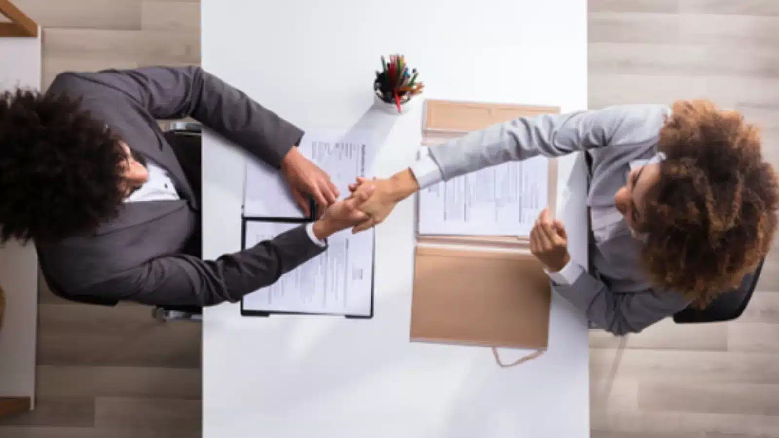 Dos personas se dan la mano sobre una mesa de despacho con currículums, vistas desde arriba